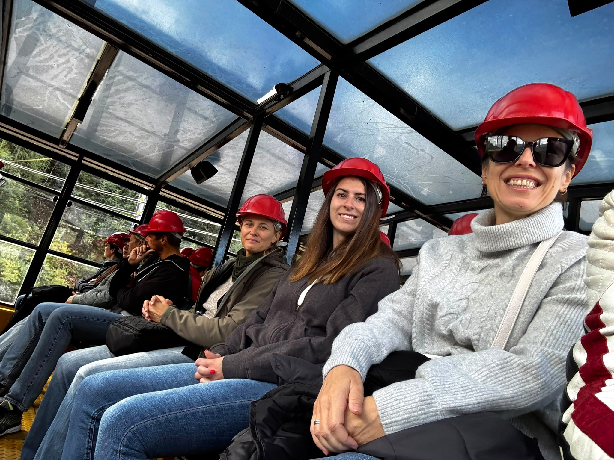 Sechs Personen mit roten Schutzhelmen sitzen in einer Gondel mit Glasdach und blicken in verschiedene Richtungen.