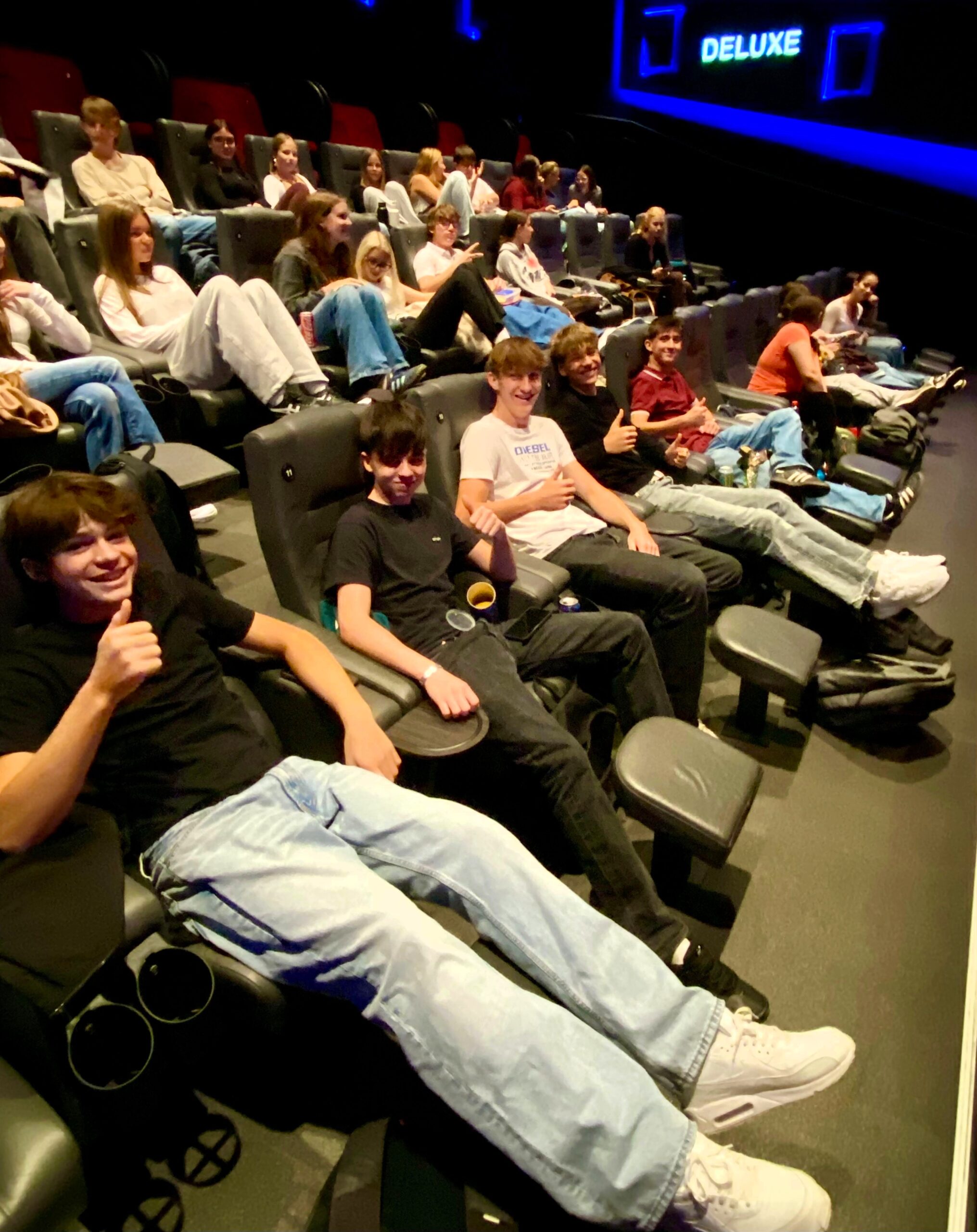 Jugendliche im Kino, einige gestikulieren und lächeln, Kinosaal mit DELUXE-Schriftzug